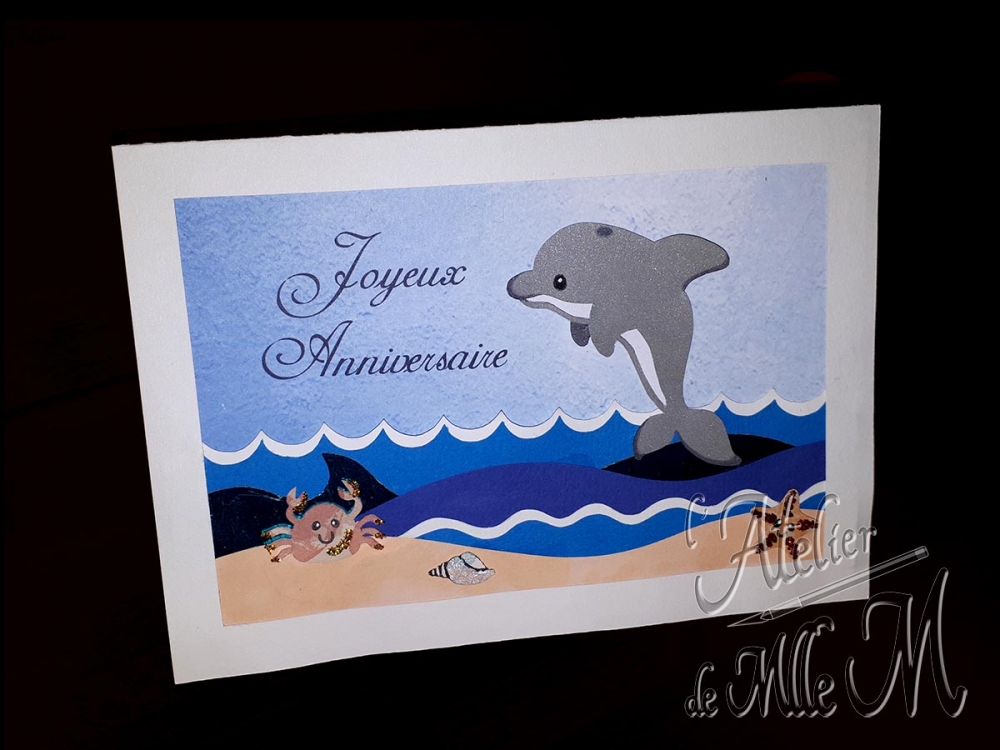 Une jolie carte d'anniversaire personnalisée. La superposition de plusieurs papiers de couleurs différente donne un effet de profondeur à la mer, et le dauphin semble sauter dans les airs grâce au scotch double face mousse. Composition : Papier de différentes couleurs et différents grammages – Paillettes.