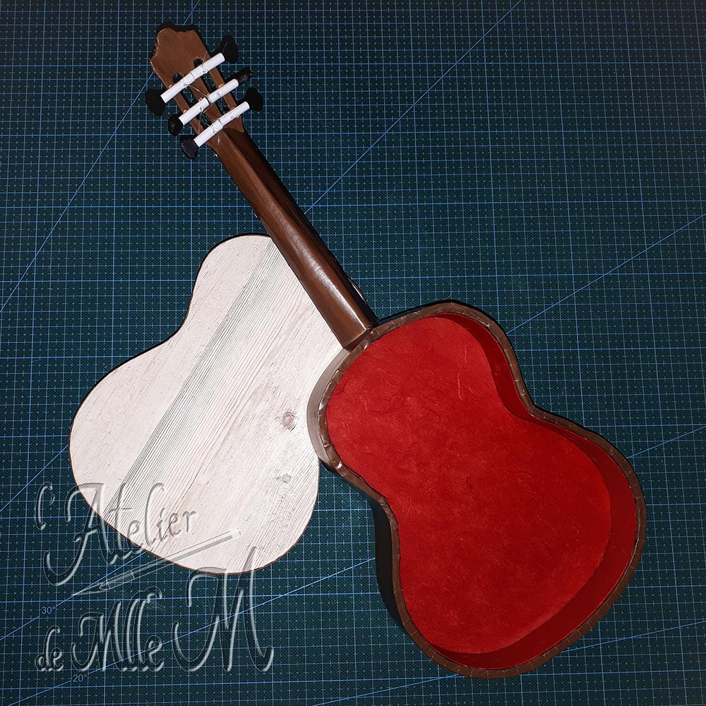 Une belle boîte en forme de guitare. Ne dirait-on pas une vraie ?! Composition : Carton. Papiers de décoration et imitation bois. Cure-dents – Fils de nylon. Dimensions : 10 (H) x 40 (L) x 15 (l) cm.