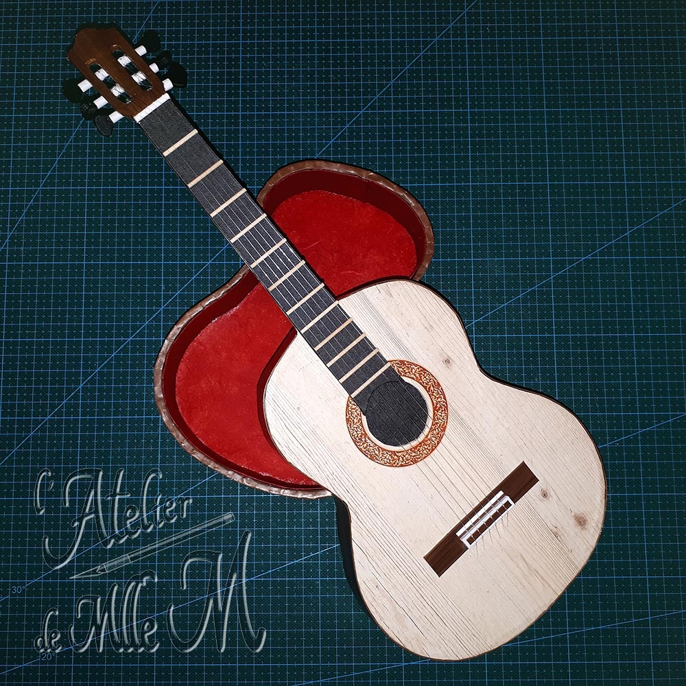 Une belle boîte en forme de guitare. Ne dirait-on pas une vraie ?! Composition : Carton. Papiers de décoration et imitation bois. Cure-dents – Fils de nylon. Dimensions : 10 (H) x 40 (L) x 15 (l) cm.